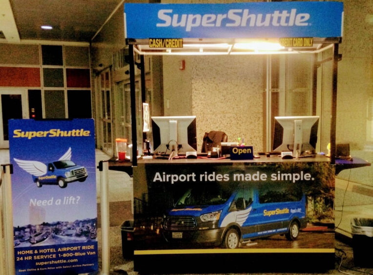 Branded SuperShuttle airport transit retail kiosk with overhead canopy and display screens.